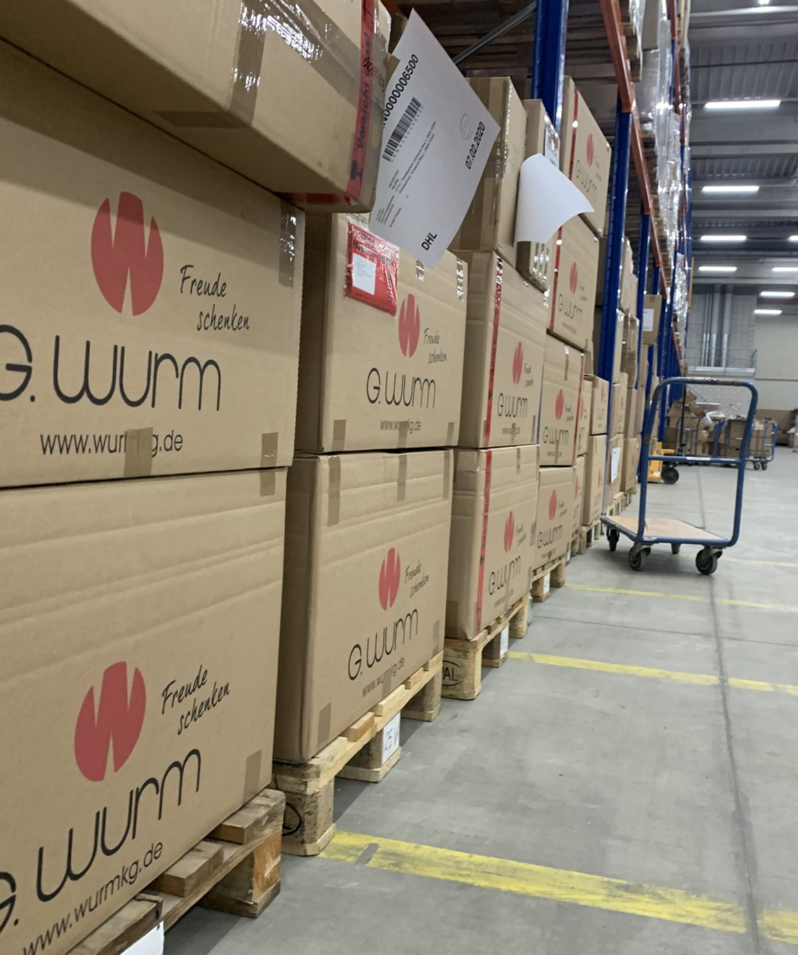 Warehouse aisles with stacks of G.wurm cardboard boxes on wooden pallets and a empty flat cart.