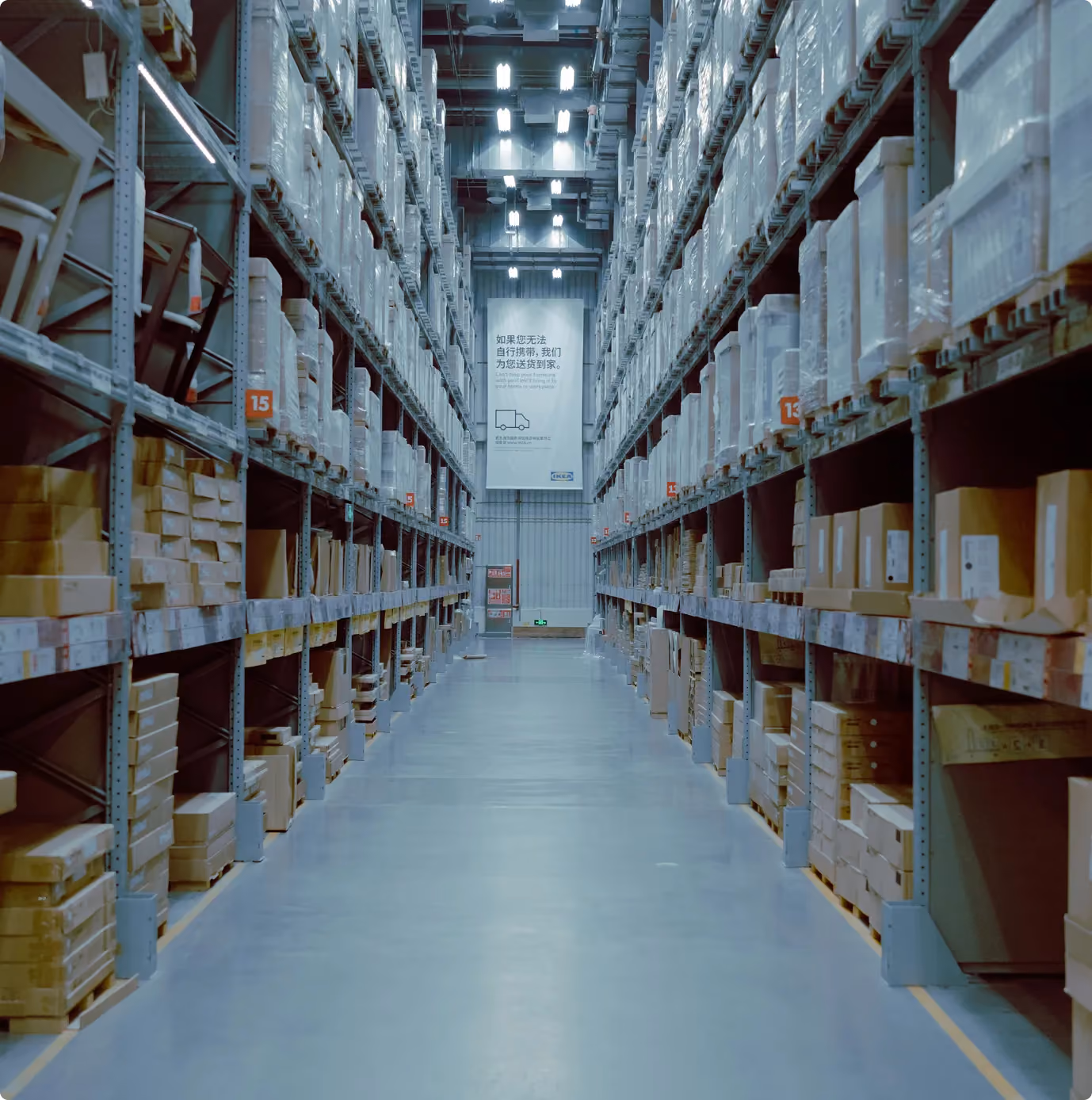 A wide aisle in a warehouse with tall shelves stocked with boxes and wrapped items, sign at center back.