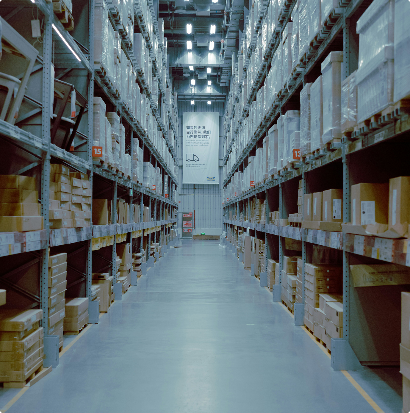 A wide aisle in a warehouse with tall shelves stocked with boxes and wrapped items, sign at center back.
