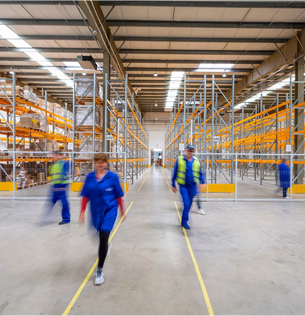 Warehouse interior with shelves stocked on left and workers walking along the central aisle.