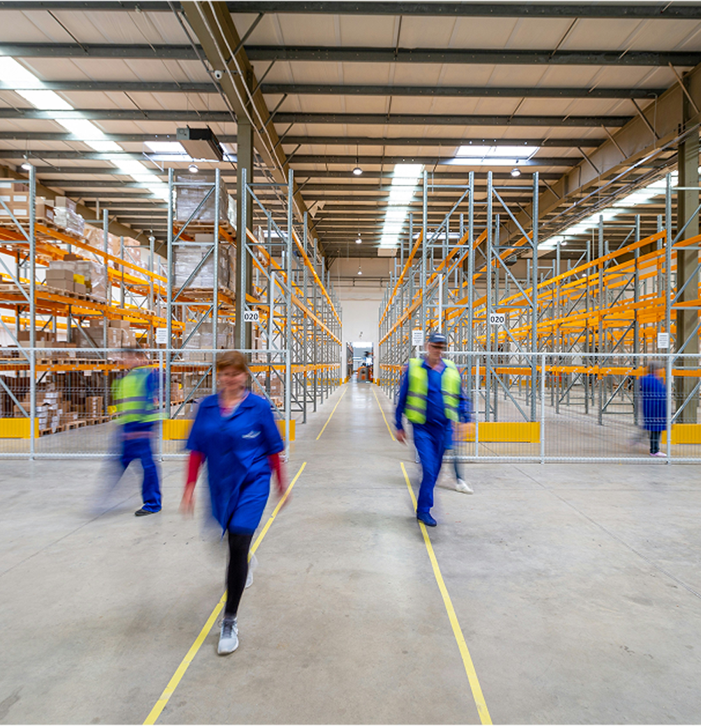 Warehouse interior with shelves stocked on left and workers walking along the central aisle.