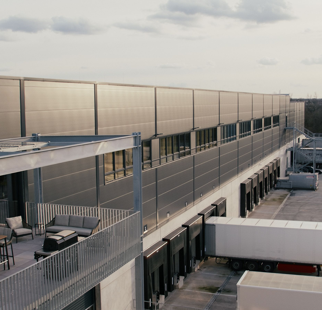 Loading docks with trucks parked alongside a large industrial warehouse building and an adjacent outdoor seating area.