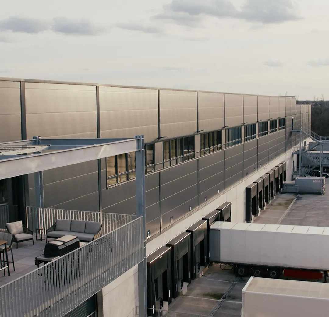 Loading docks with trucks parked alongside a large industrial warehouse building and an adjacent outdoor seating area.