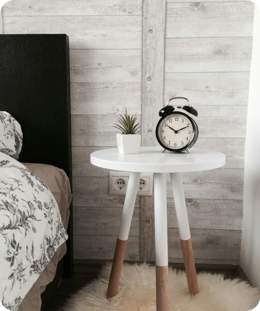 White bedside table with wooden legs holding a black alarm clock and small potted plant next to a bed.