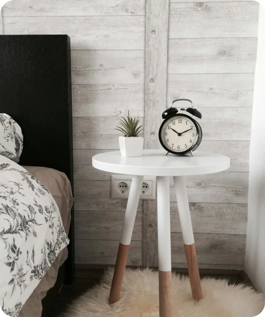 White bedside table with wooden legs holding a black alarm clock and small potted plant next to a bed.