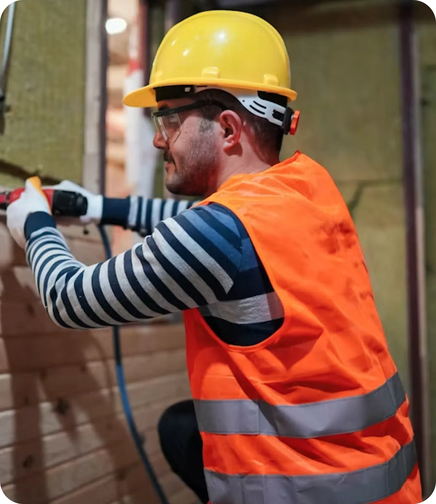 Technician in a yellow hard hat, safety glasses and orange hi-vis vest using a power tool on a timber wall.