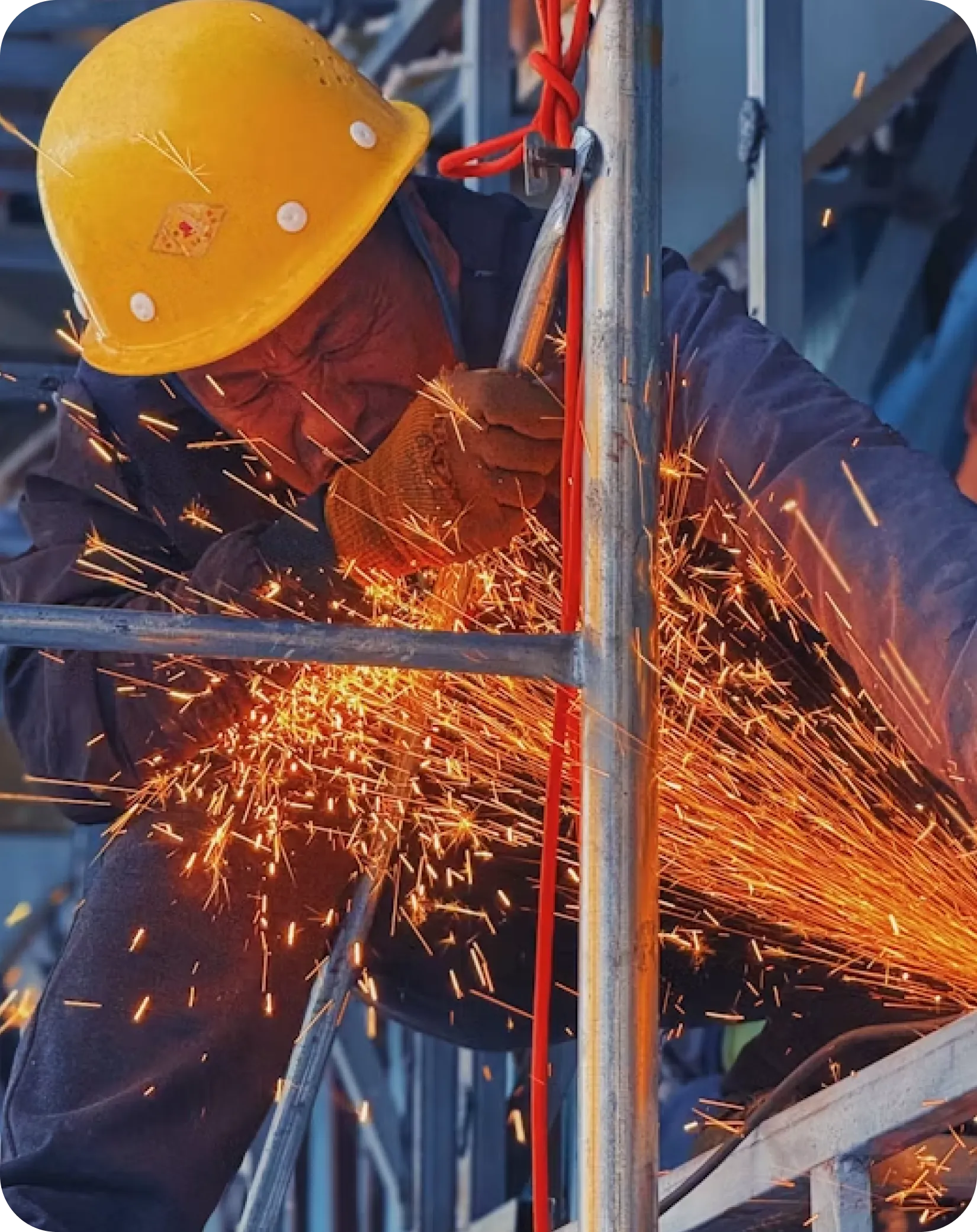 Worker in a yellow hard hat cutting metal scaffolding with an angle grinder as sparks fly.