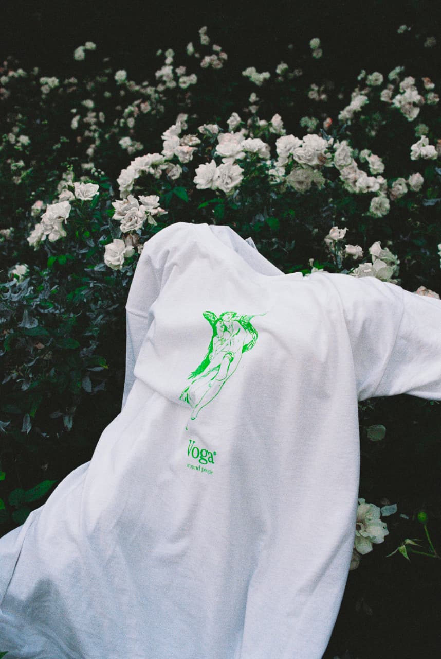 White T-shirt with green illustration of two embracing figures and text 'Voga' laid over dark green foliage and white flowers.