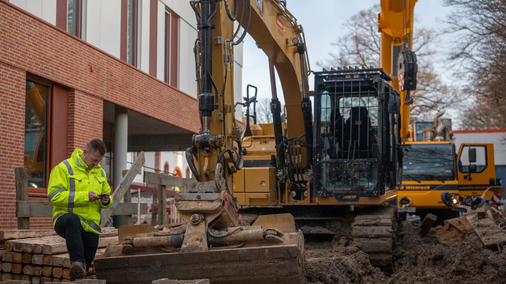 Eine Person in einer leuchtend gelben Sicherheitsjacke steht auf einem Holzstapel und blickt auf ein Smartphone neben einem großen gelben Bagger auf einer Baustelle.