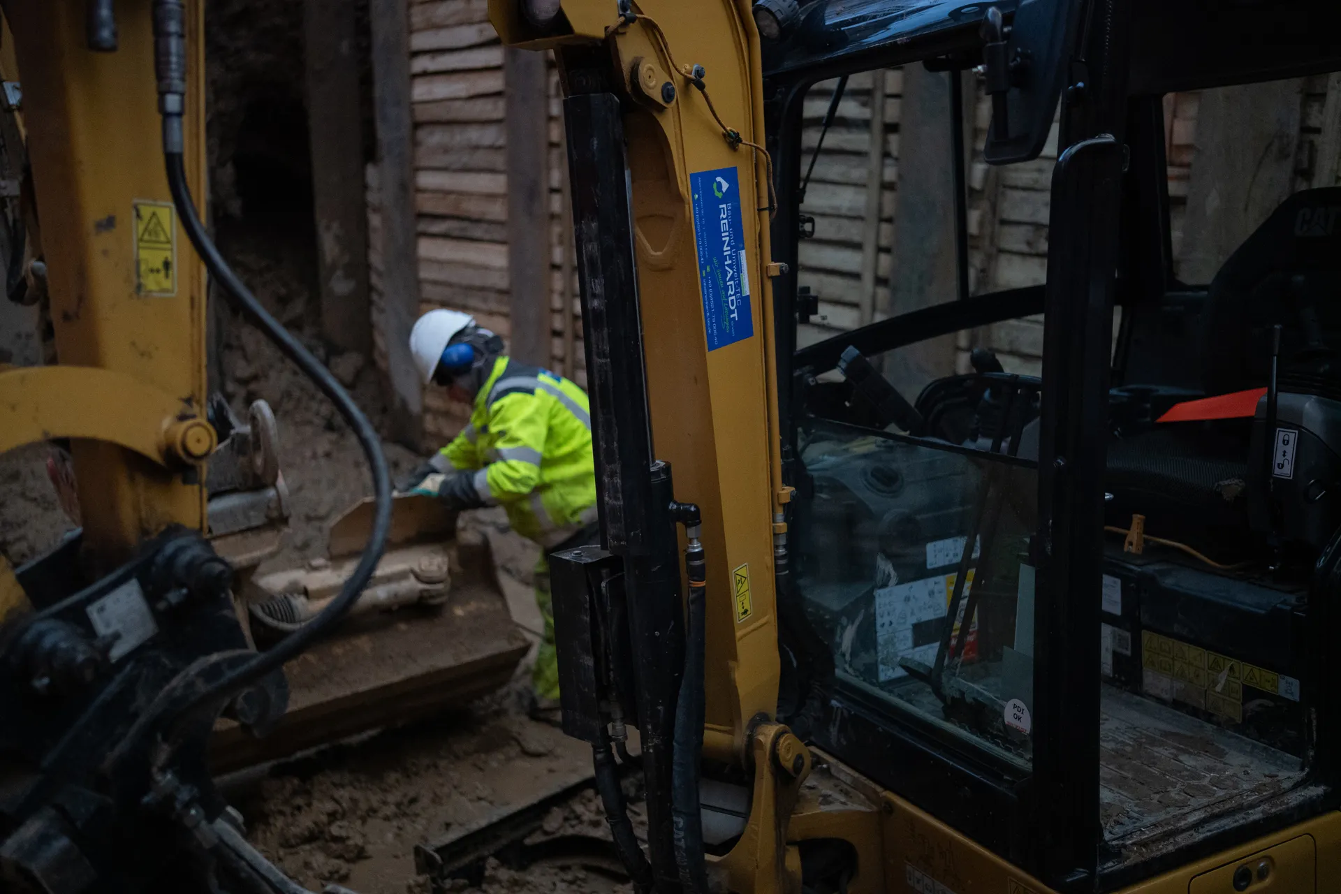 Bauarbeiter in gelber Schutzkleidung und Helm arbeitet neben einem gelben Bagger auf einer schlammigen Baustelle.