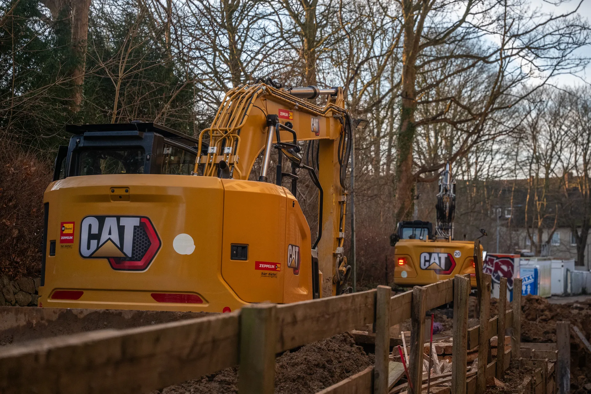 Zwei gelbe CAT-Bagger stehen auf einer Baustelle mit Erde und Holzzäunen, im Hintergrund Bäume ohne Blätter.