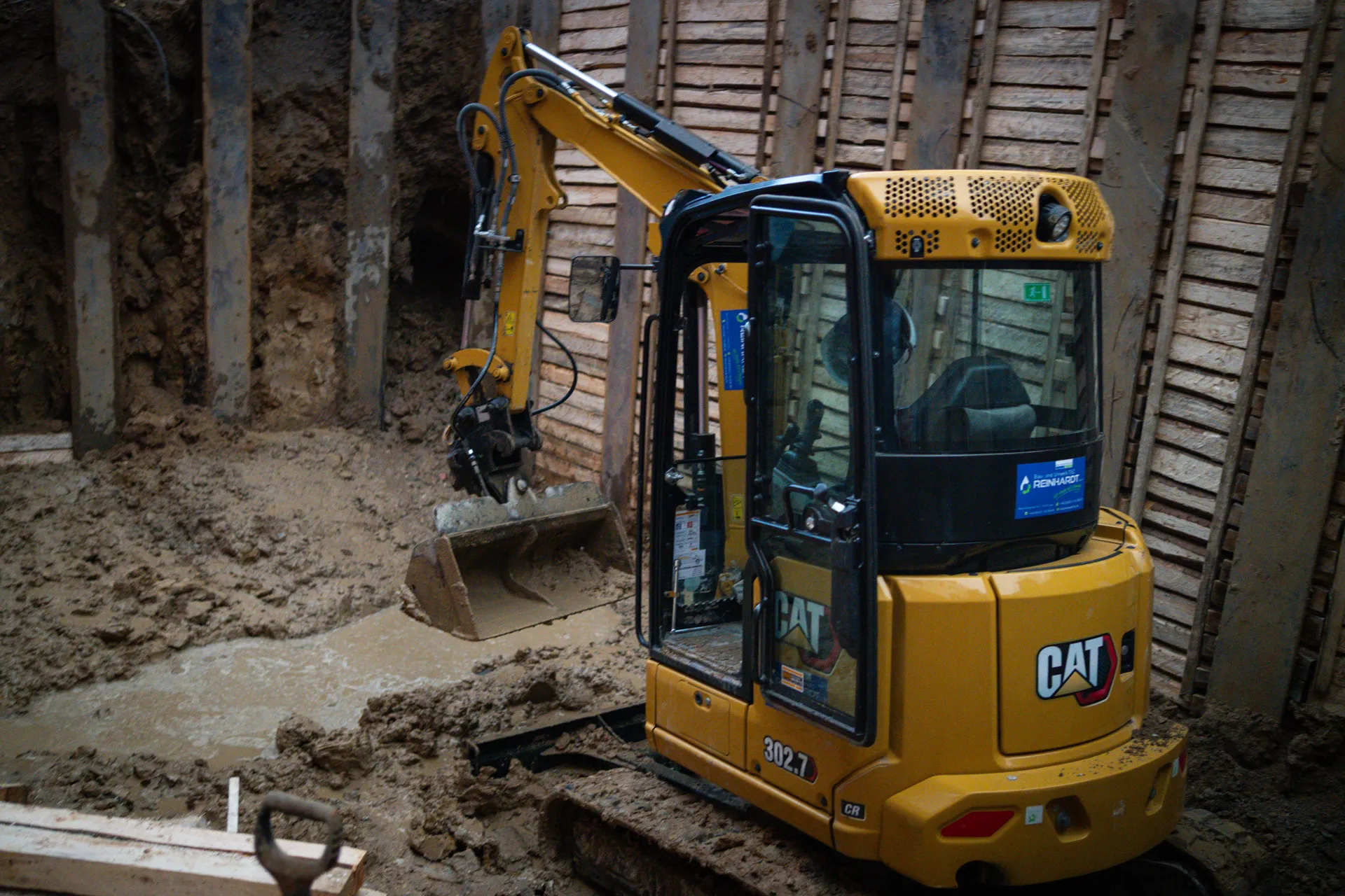 Gelber CAT-Bagger in einer schlammigen Grube mit Holzwandstützen.