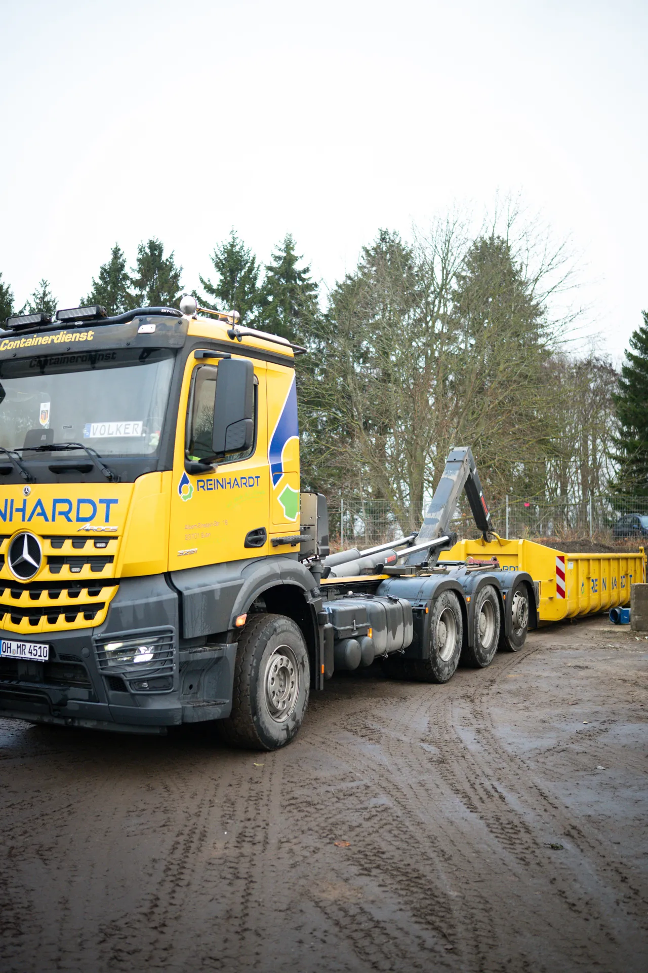 Gelber Reinhardt LKW mit Container auf schlammigem Baustellengelände und kahlen Bäumen im Hintergrund.