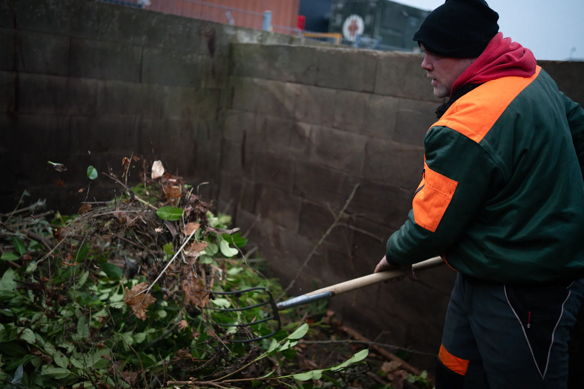 Mann in Arbeitskleidung hebt Gartenabfälle mit einer Mistgabel auf.