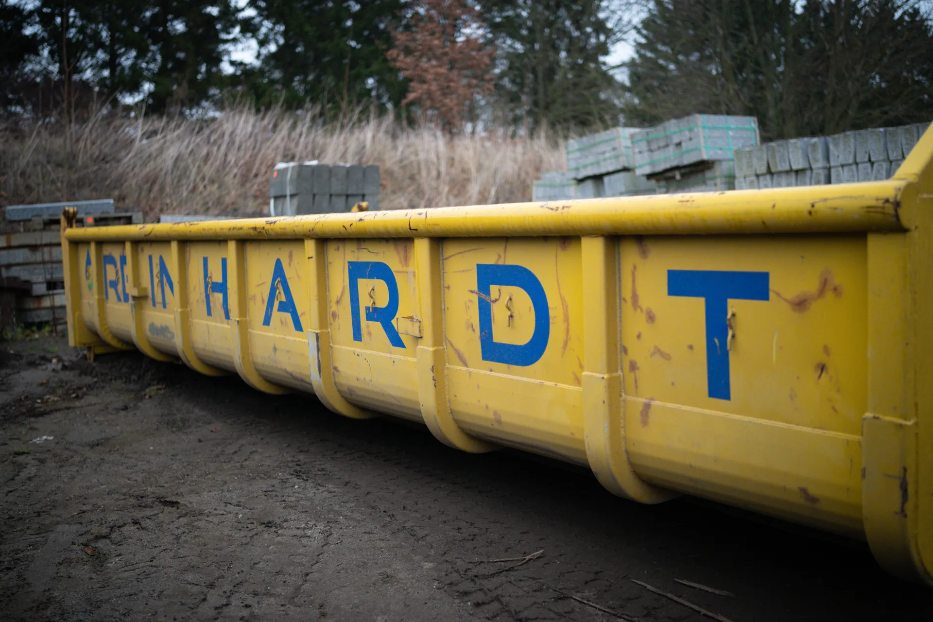Gelber Müllcontainer mit blauer Aufschrift 'REINHARDT' auf einem dirtigen Boden im Freien.