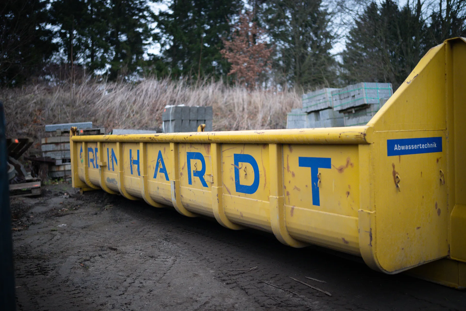 Gelber Abfallcontainer mit der Aufschrift 'REINHARDT' und einem Aufkleber 'Abwassertechnik' in einer Industrieumgebung.