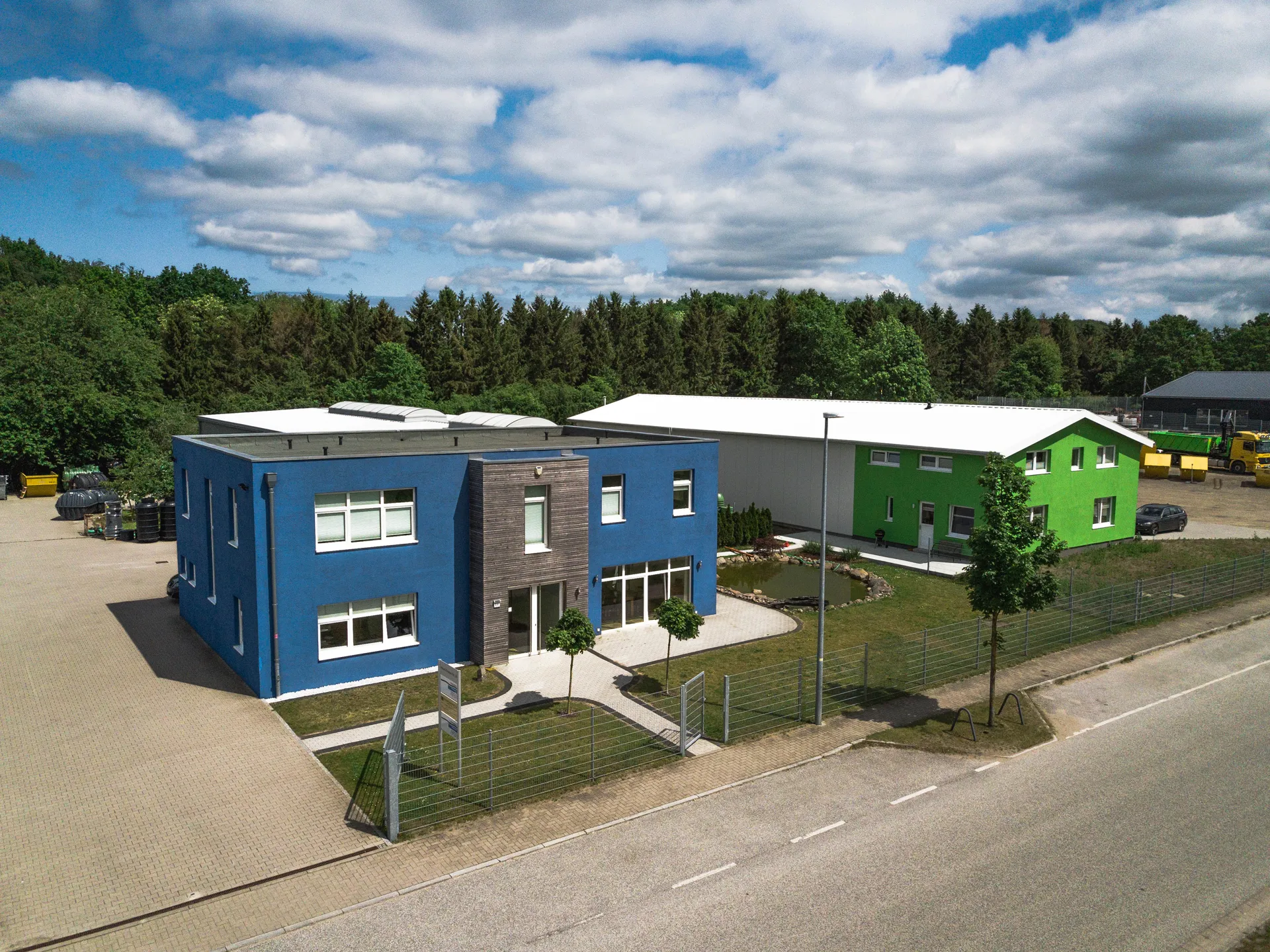 Blick auf zwei moderne Firmengebäude, eines blau und das andere grün, umgeben von Grünflächen und einem kleinen Teich, mit bewölktem Himmel und Wald im Hintergrund.