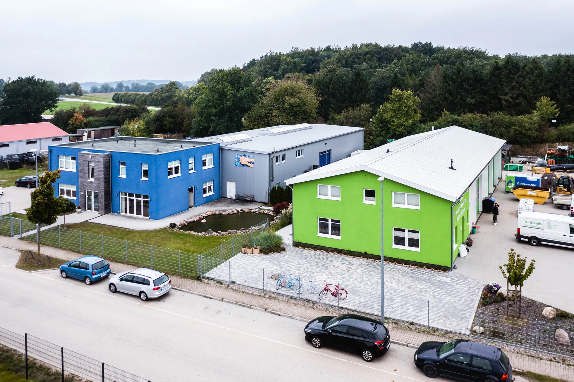 Luftaufnahme eines modernen Büro- und Industriekomplexes mit blauem und grünem Gebäude, einem Teich und geparkten Autos entlang der Straße.