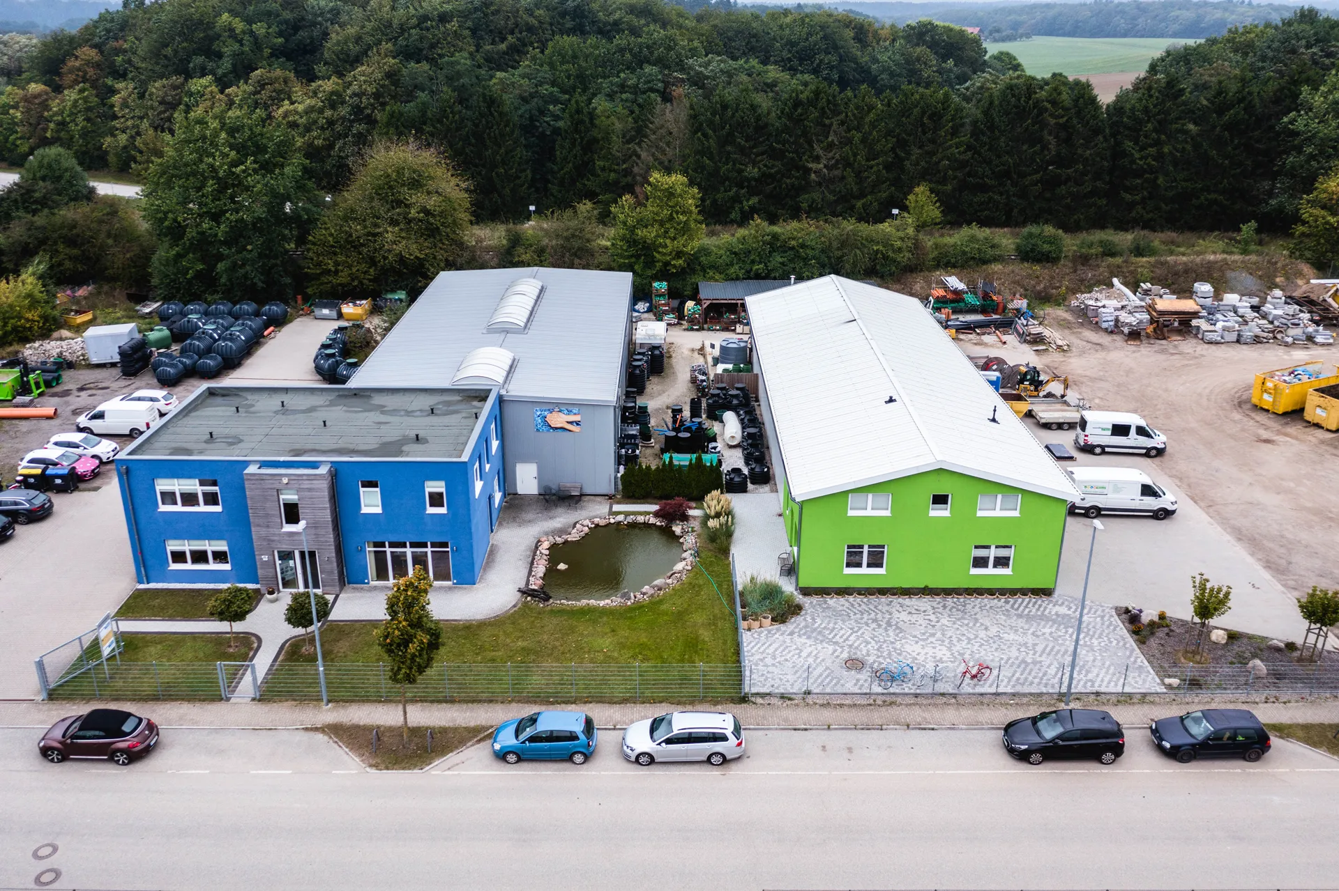 Luftaufnahme von zwei nebeneinanderliegenden Fabrikgebäuden, eines blau und eines grün, mit Parkplatz, Bäumen und einem kleinen Teich dazwischen.