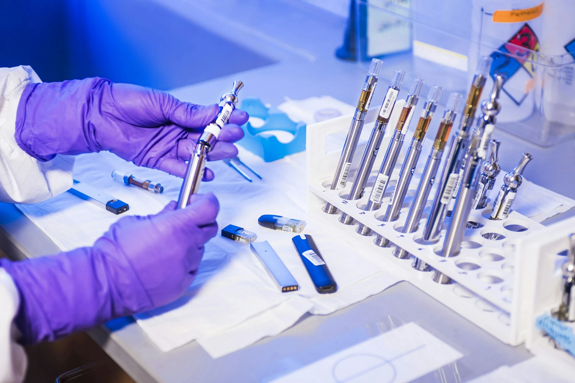 Hands wearing purple gloves holding a vape pen next to a rack of multiple vape cartridges on a lab table.