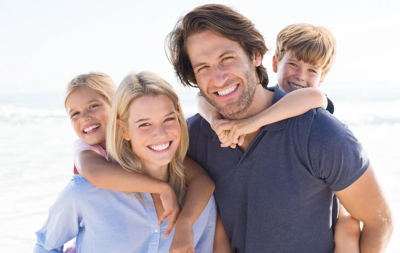 Smiling family of four posing at the beach with children hugging parents from behind.