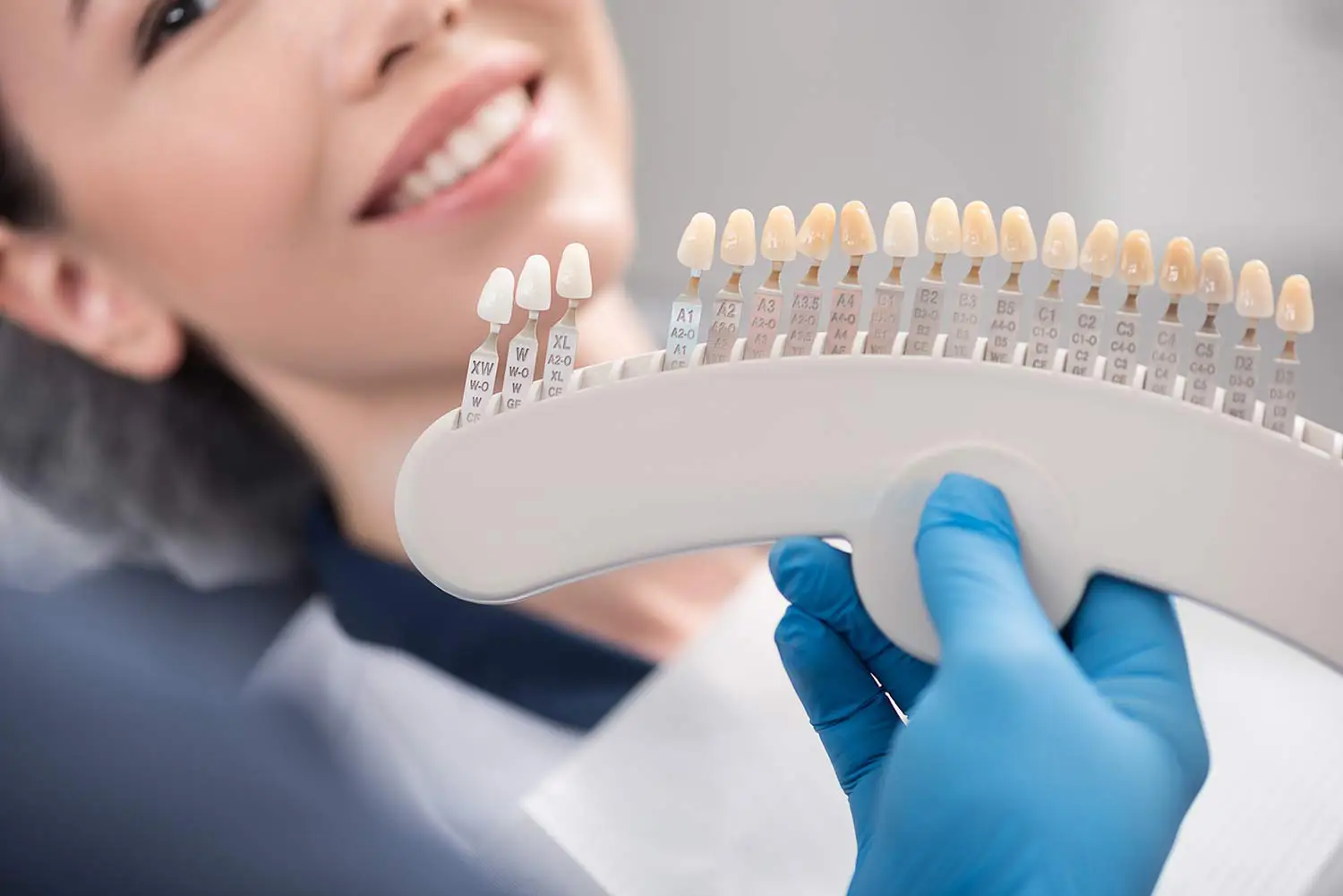 Dentist in blue gloves holding a dental shade guide near a smiling patient's mouth.