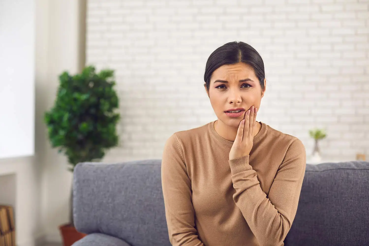 Woman sitting on a couch with a pained expression, holding her cheek as if experiencing toothache.