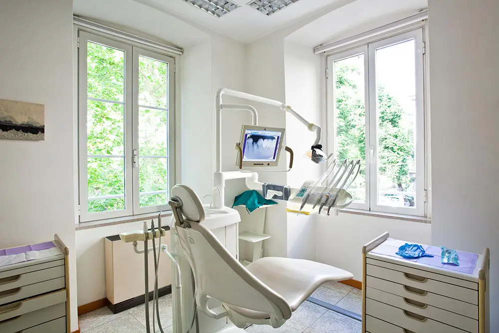 Bright dental clinic room with a dental chair, X-ray screen showing teeth, and large windows with green trees outside.
