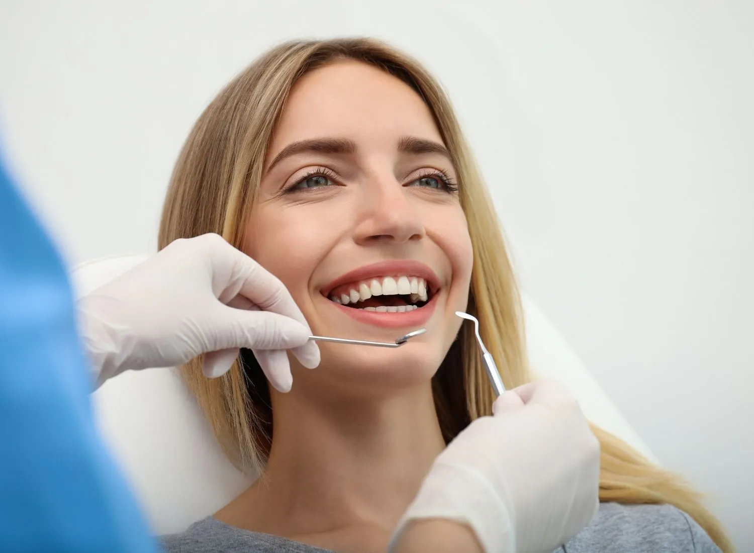 Dentist wearing gloves examining smiling woman's teeth with dental tools in a clinic.