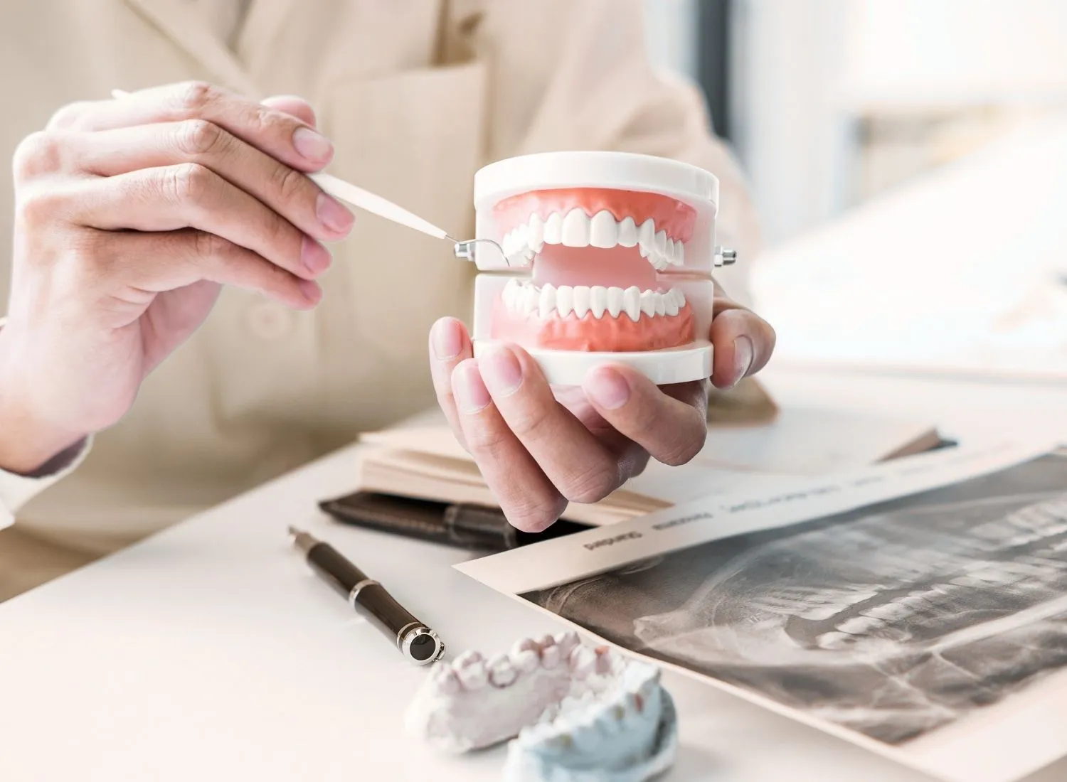 Hands holding a dental model and tool with X-ray and tooth molds on a white table.