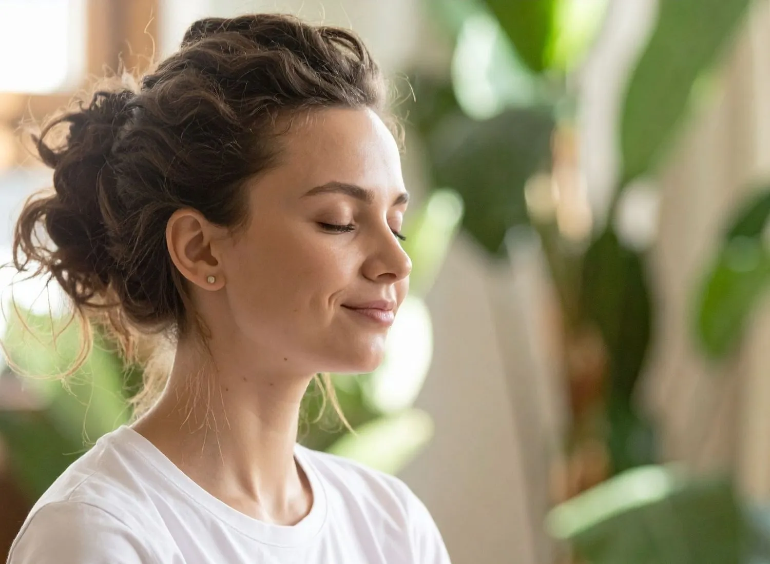 Woman with eyes closed and hair tied back, smiling gently with indoor green plants in background.