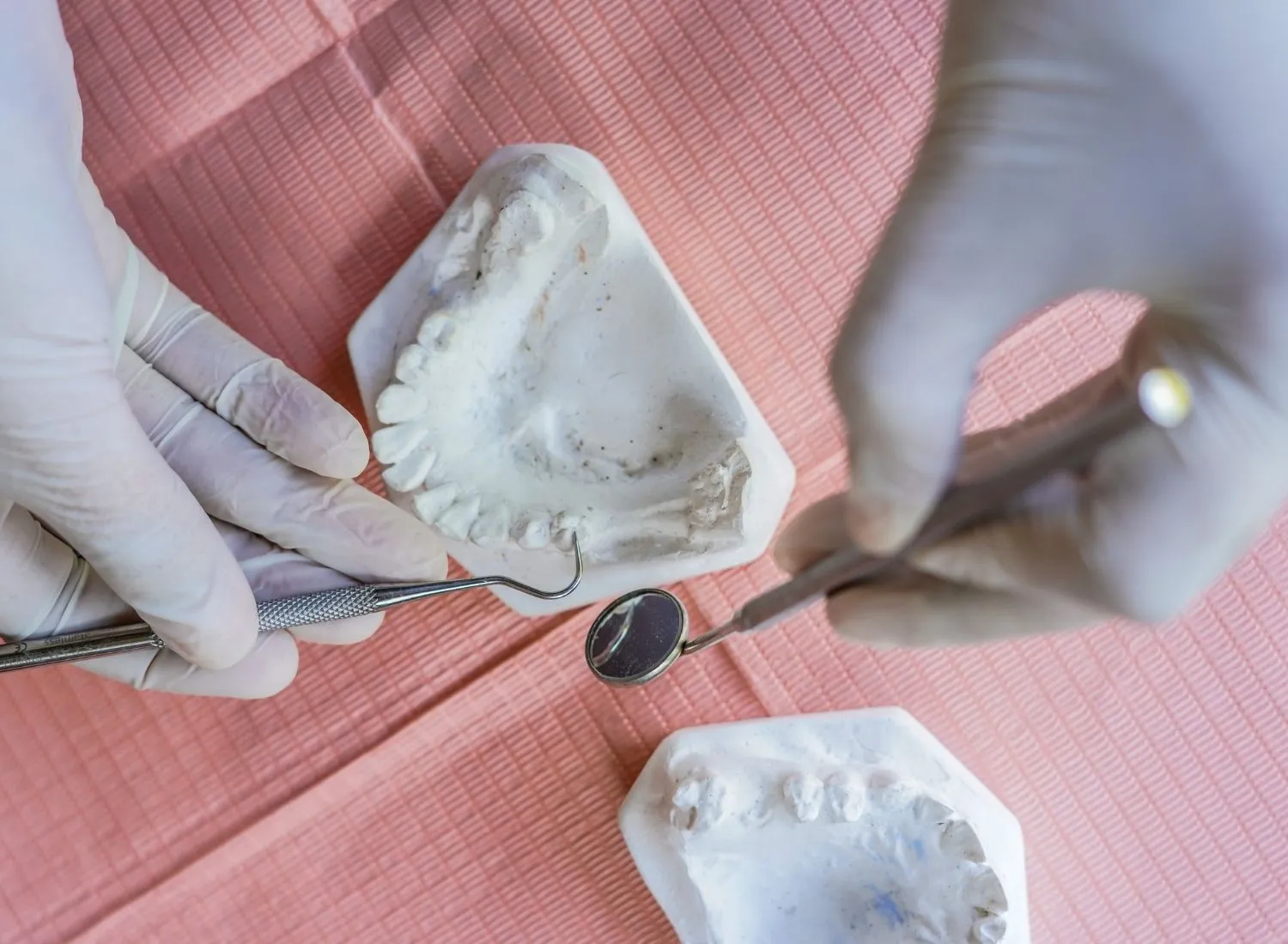 Hands in gloves holding dental tools near plaster dental molds on a pink surface.