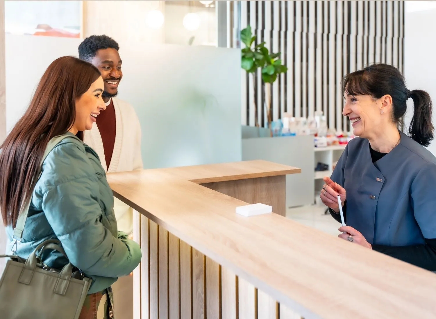 Smiling woman and man talking to a female receptionist at a modern clinic front desk.