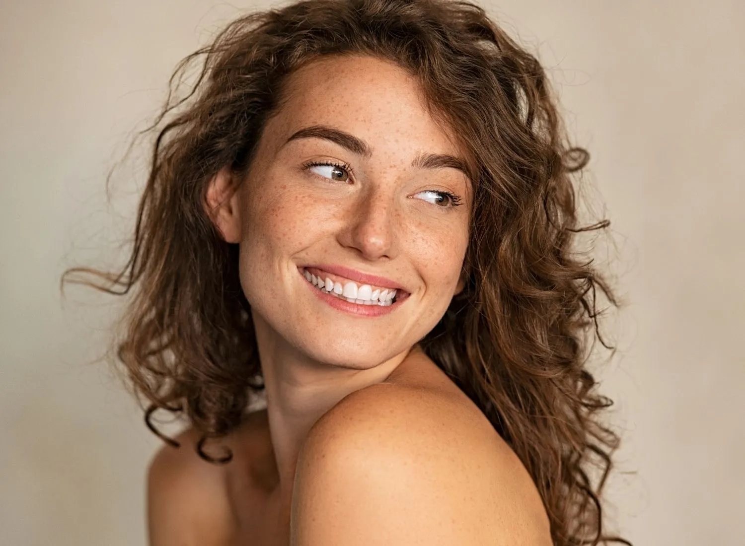 Smiling woman with curly hair and freckles looking to the side against a neutral background.
