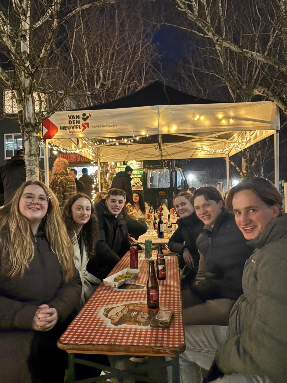 Groep jongeren zittend aan een lange tafel met drankjes onder een verlicht feesttent in de avond.
