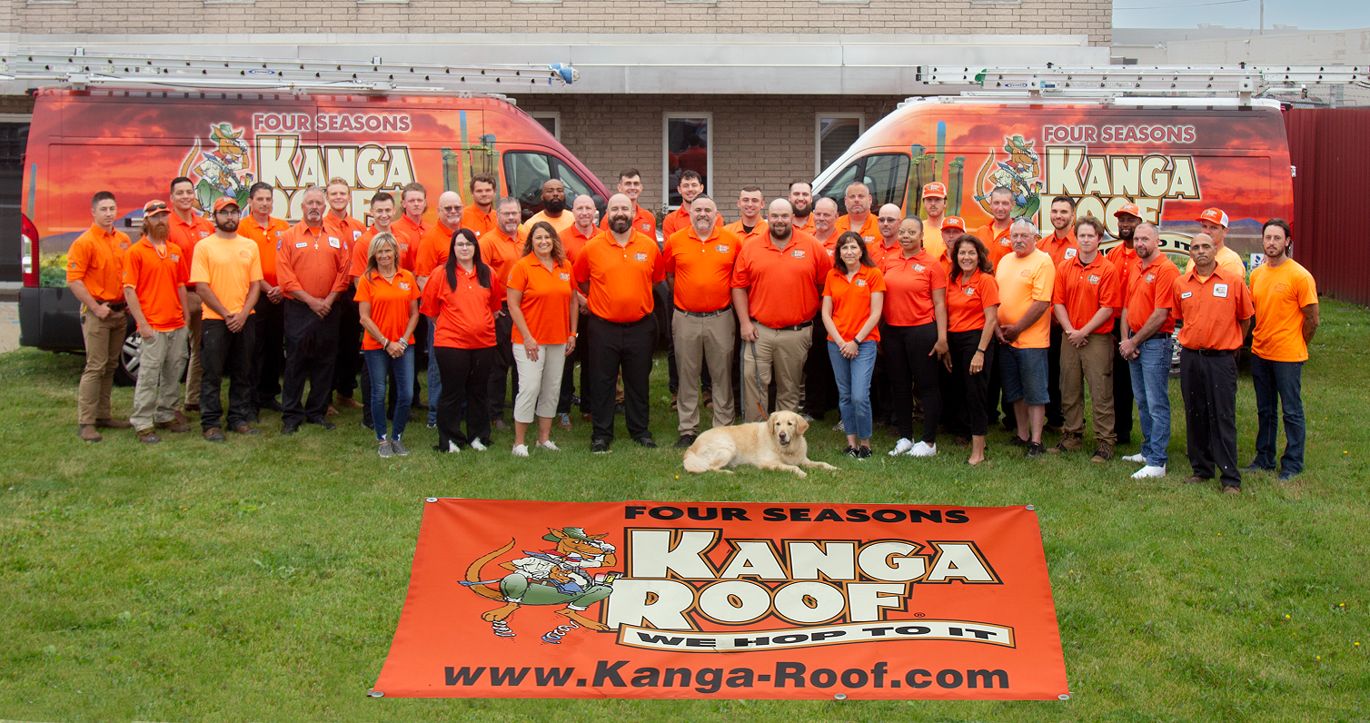 Group of workers in orange shirts posing in front of Kanga Roof vans and a banner on the grass.