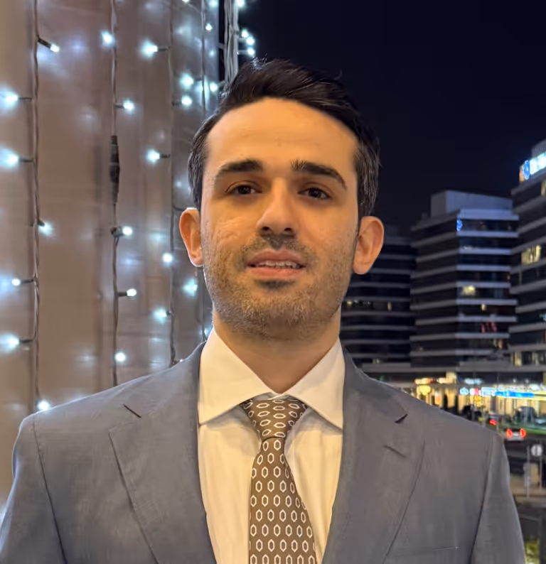 Man wearing a grey suit and patterned tie standing outdoors at night with lit buildings and string lights in the background.