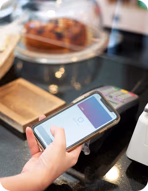 Hand holding a smartphone displaying a digital payment app near a credit card terminal at a store counter.