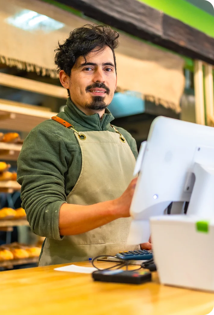 Man wearing a green sweater and beige apron standing behind a bakery counter operating a payment terminal.