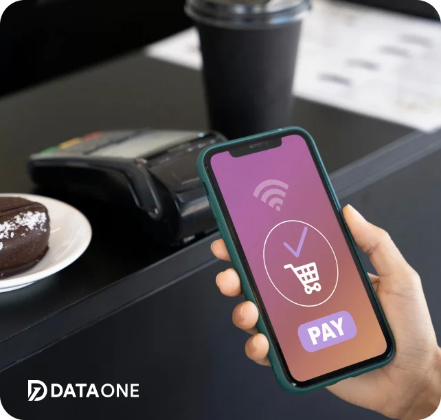 Hand holding smartphone displaying a digital payment screen with a shopping cart icon and 'PAY' button, next to a card reader and a plate with a chocolate dessert.