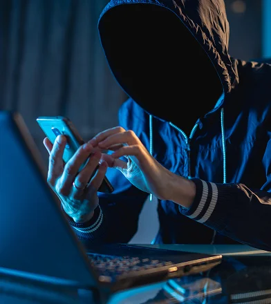 Person in hoodie using smartphone with laptop in dark room.