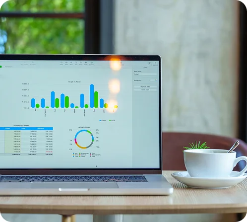 Laptop on a wooden table displaying a budget report with graphs and charts, next to a white cup with a small plant and spoon on a saucer.