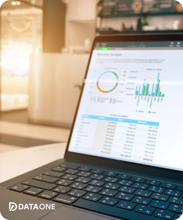 Laptop displaying a monthly budget dashboard with charts and tables, placed on a white surface in a bright room.