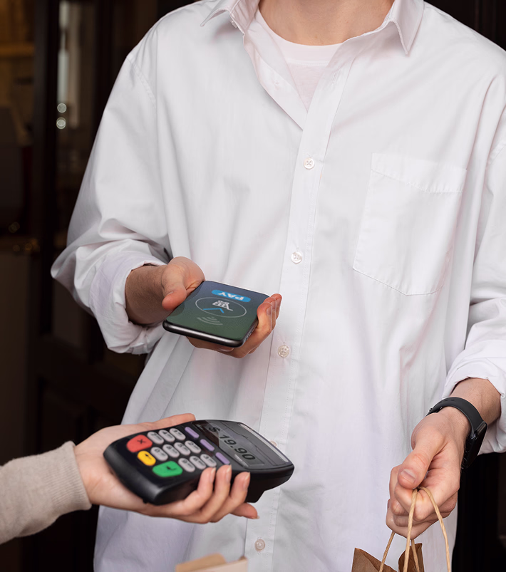 Person in a white shirt holding a smartphone near a payment terminal for contactless payment while carrying paper bags.