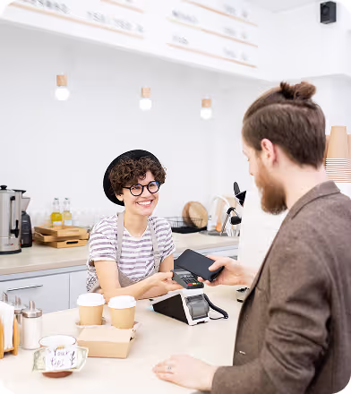 Barista smiling while processing a contactless payment from a customer at a coffee shop counter.