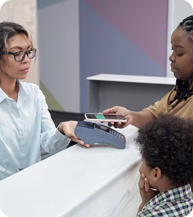A woman uses a smartphone to make a contactless payment at a counter while a child watches.