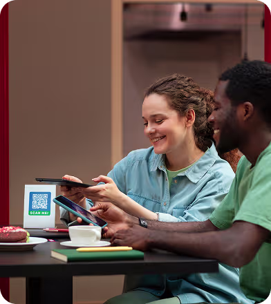 A woman scans a QR code with her smartphone at a table while a man holds the tablet displaying the code, with coffee and pastries on the table.