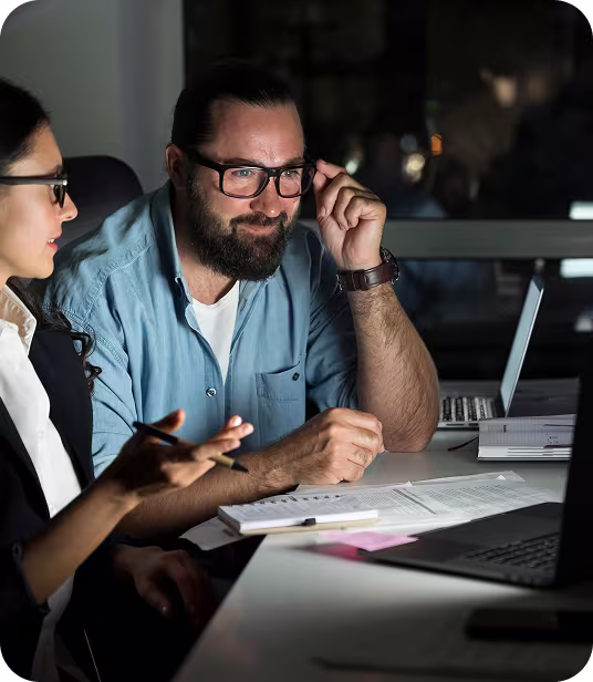Two colleagues discussing work documents at a desk in an office with laptops, one man adjusting his glasses.