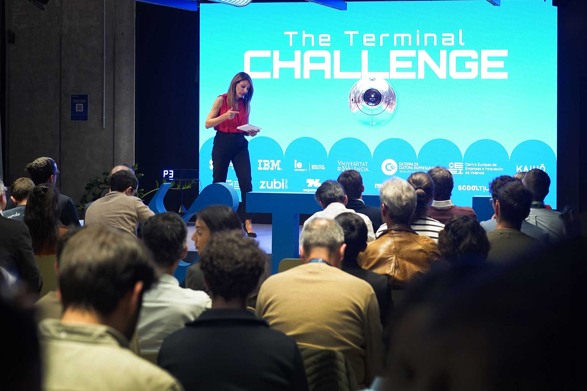 Mujer habla en escenario frente a audiencia con pantalla azul que dice The Terminal Challenge.