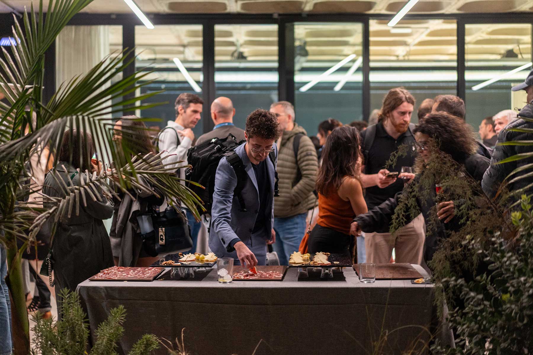 Personas en una reunión social con mesa de aperitivos y plantas decorativas alrededor.